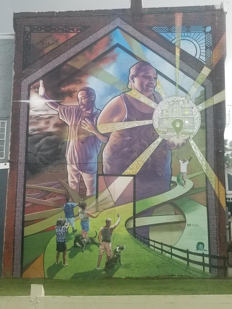 an image of a mural taking up the whole height of a building. the mural shows two people, one holding a ball of light with a building in it, the other looking into the distance. there's a path winding around these people that a group of much smaller people are beginning to walk across