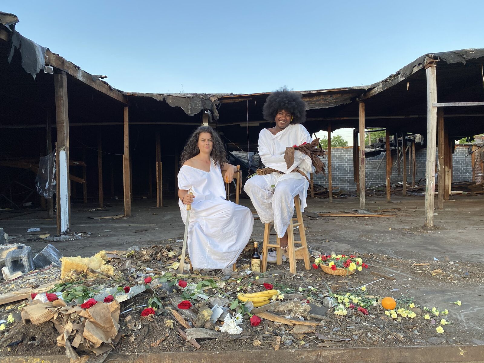 two women seated on stools in an abandoned area surrounded by flowers and food. one of them holds a machete like a walking stick