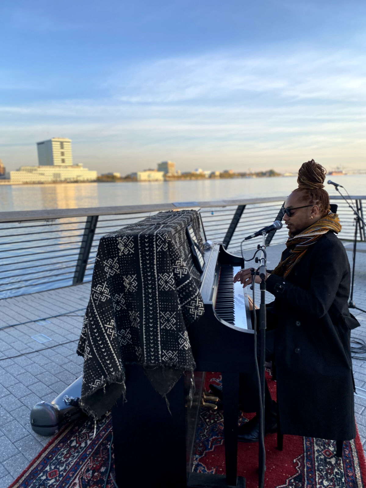man with sunglasses and dreads in an updo sings a piano in front of the waterfront