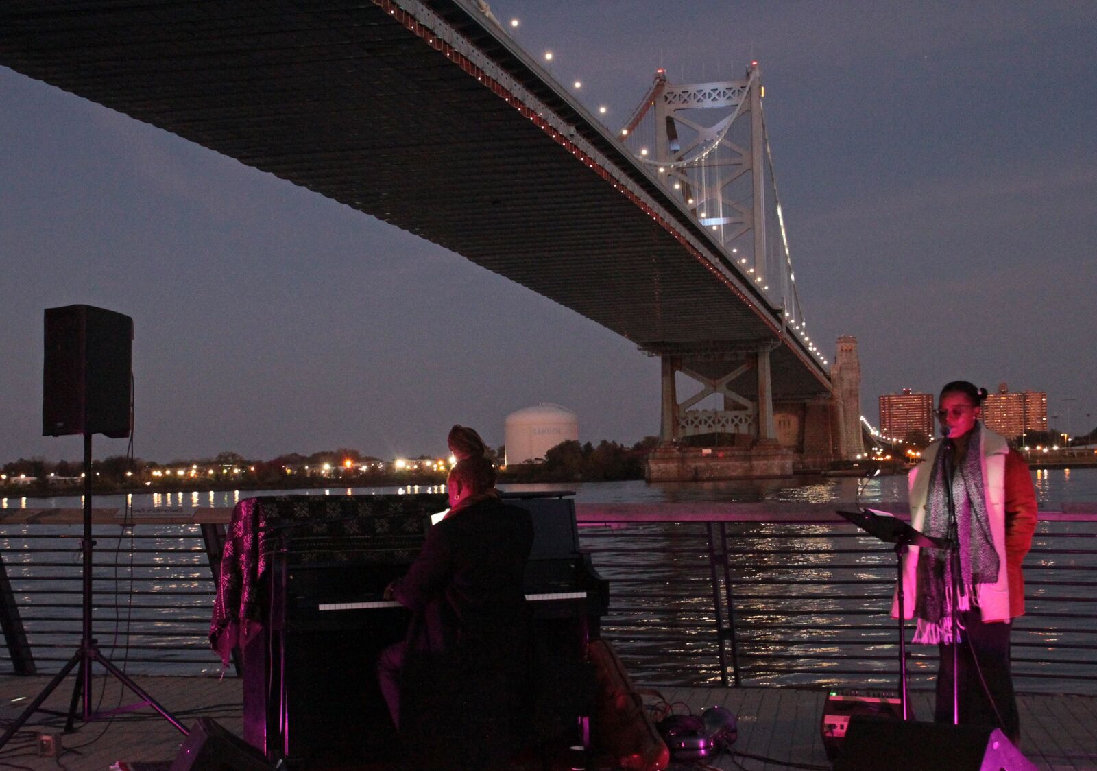 woman reads at  a music stand while man plays piano lit by red lights in front the of the waterfront and the benjamin franklin bridge at night