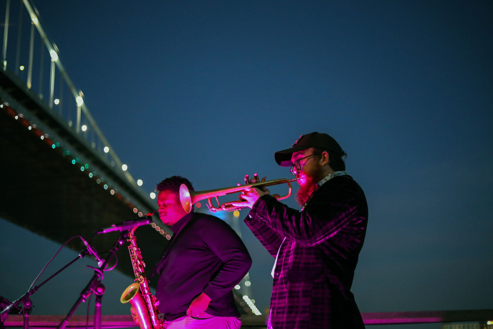 man plays trumpet and second man plays saxophone lit by red light at night in front of a bridge