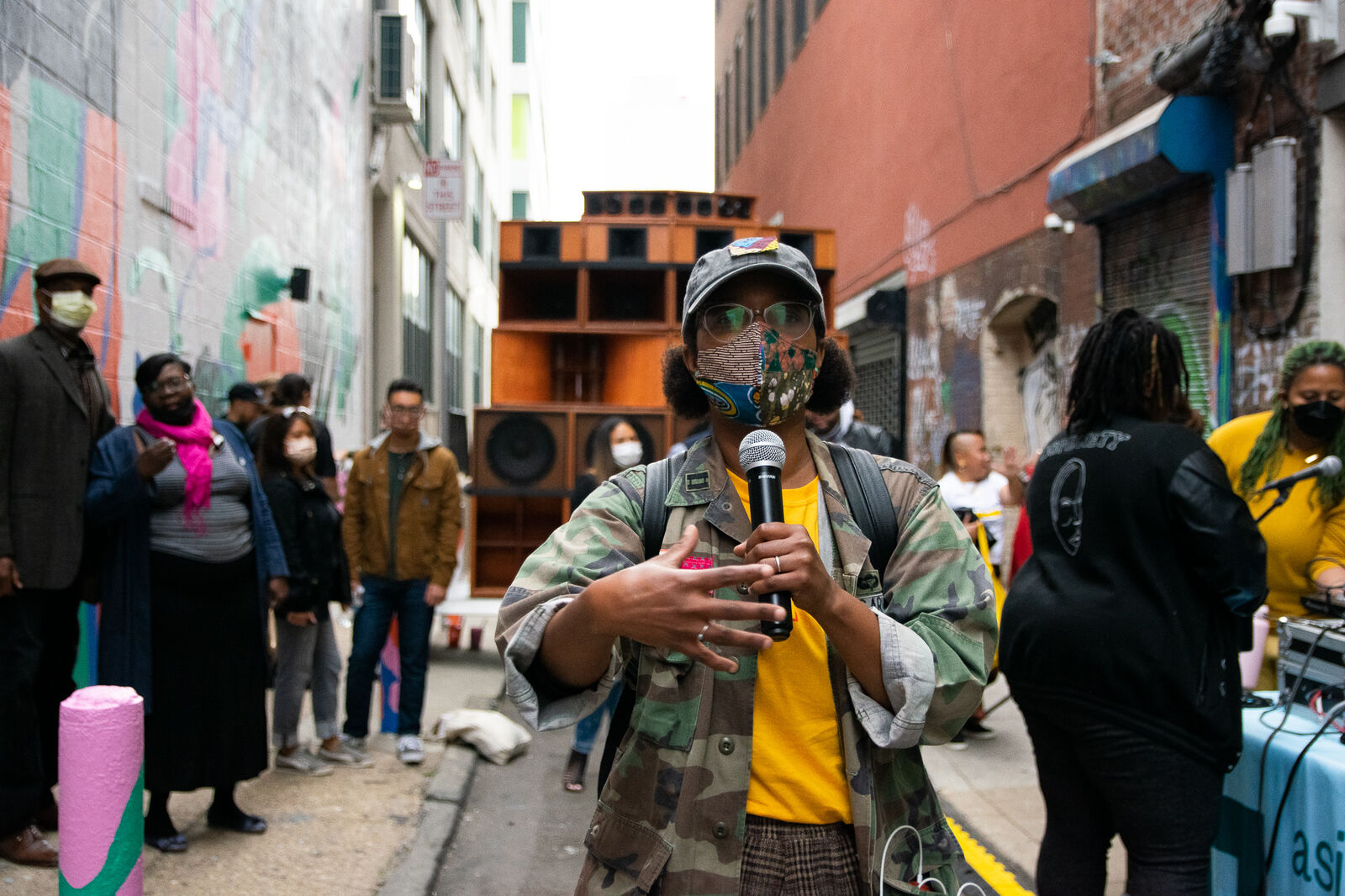 woman in mask on microphone speaks in front of a crowd and wall of speaker cabinets in the street