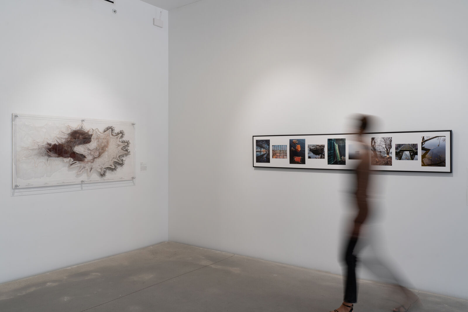 a person blurred walks in front of a collection of photography and fiber arts piece in a gallery