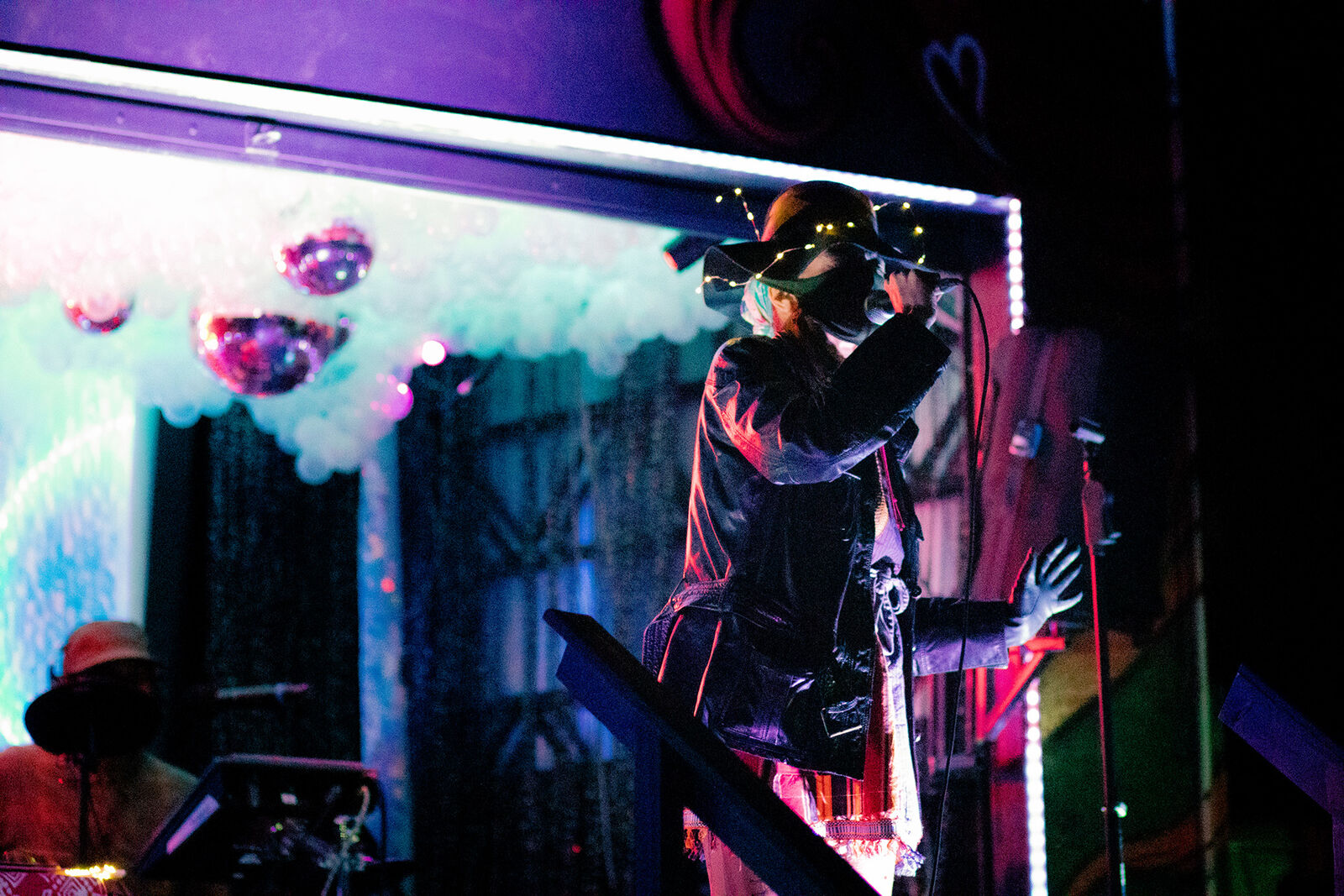 woman wearing a hat covered in twinkling lights performs in front of disco balls and pink lights