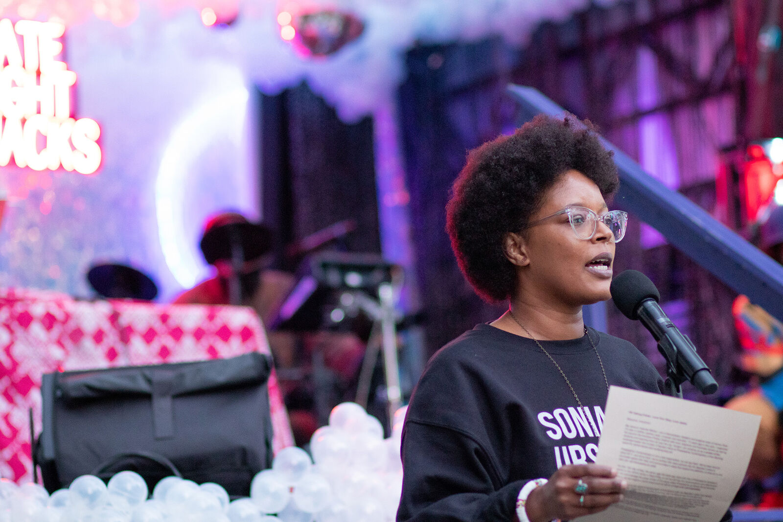 a woman speaks to a crowd in front of a pink stage with a neon sign that says "Late Night Snacks"