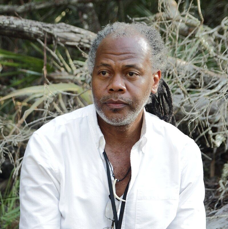 man with gray dreads in a ponytail in a white shirt stares off into the distance in front brush and trees