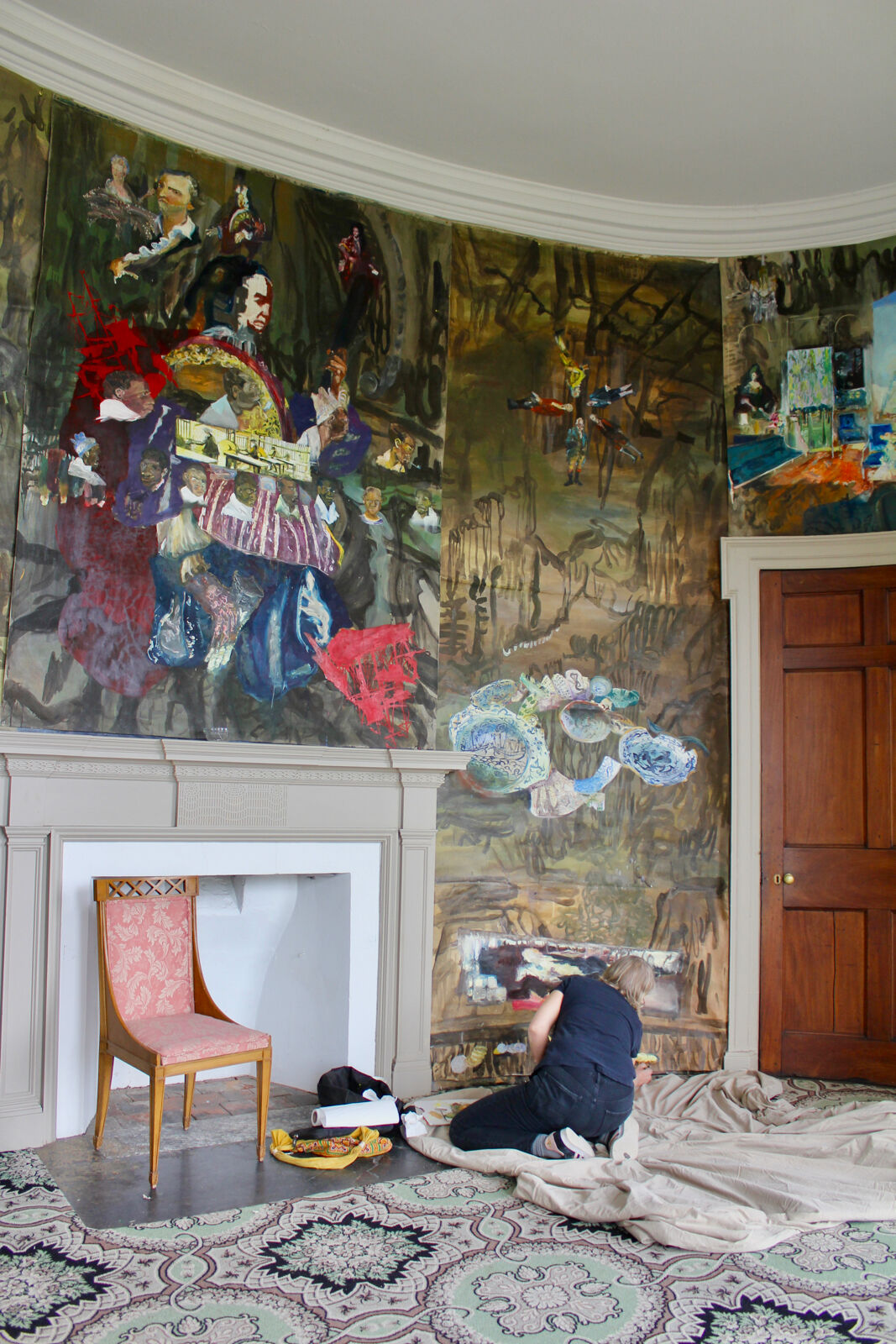 woman sit on painters cloth as she paints the walls in vibrant and expressionistic style of a historic mansion next to a fireplace and oriental rug
