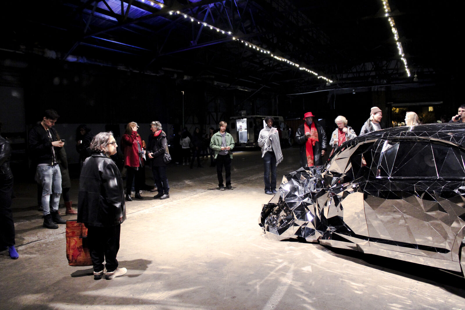 crowd gathers around sculpture of shattered car that reflects into the room