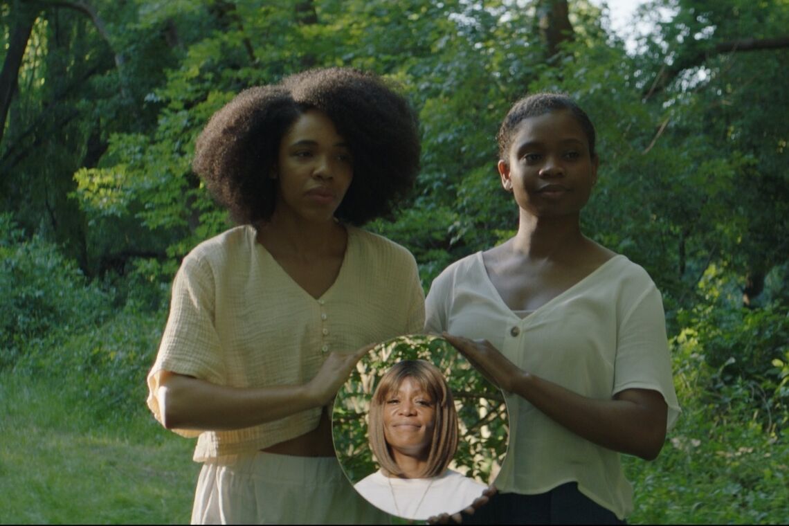two Black women hold up a circular mirror reflecting the image of another Black woman standing in front of them. All are smiling. They are standing amongst nature and trees