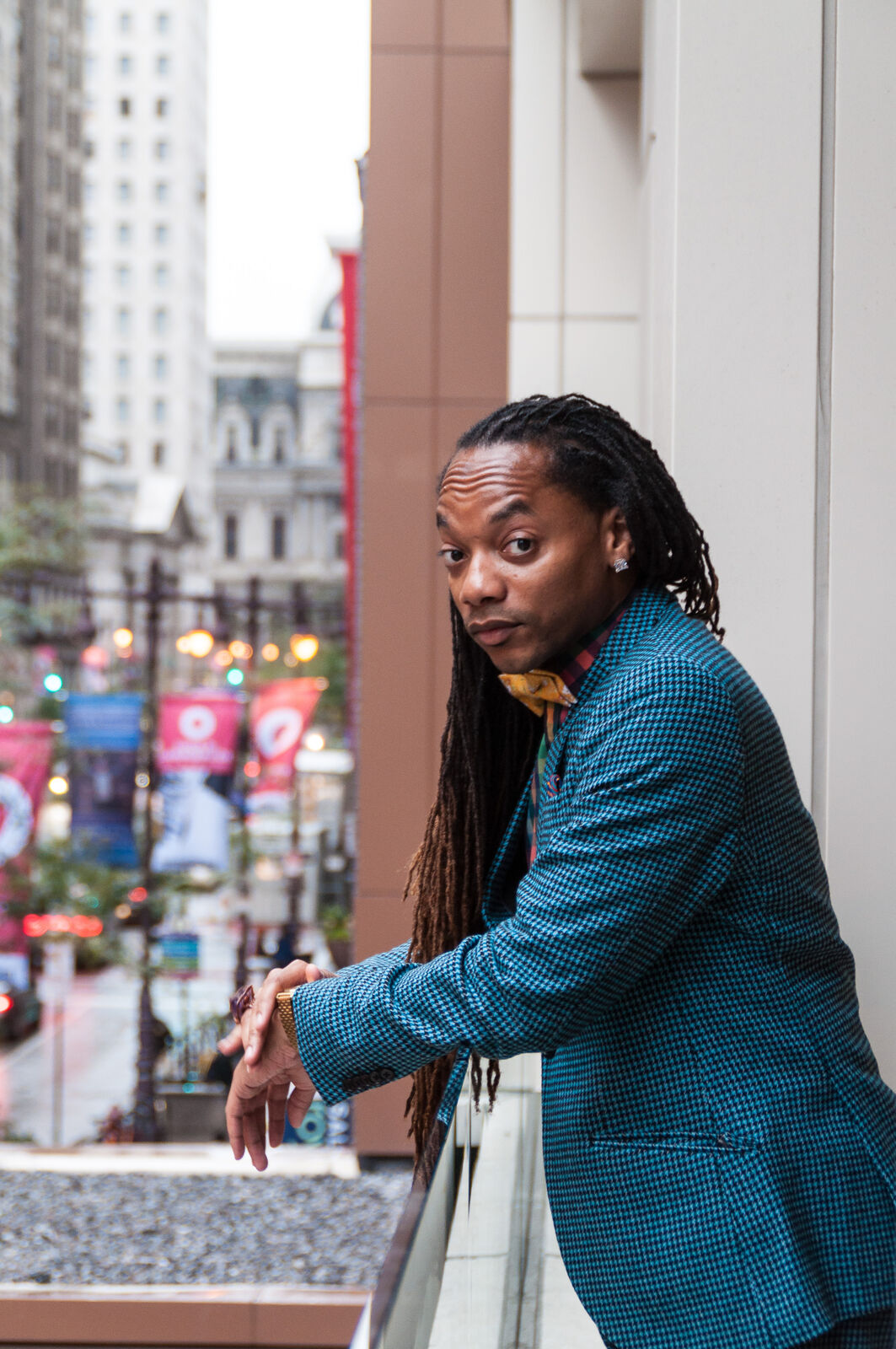 a man with long dreads stands on a balcony in front of a busy street in a suit and bowtie