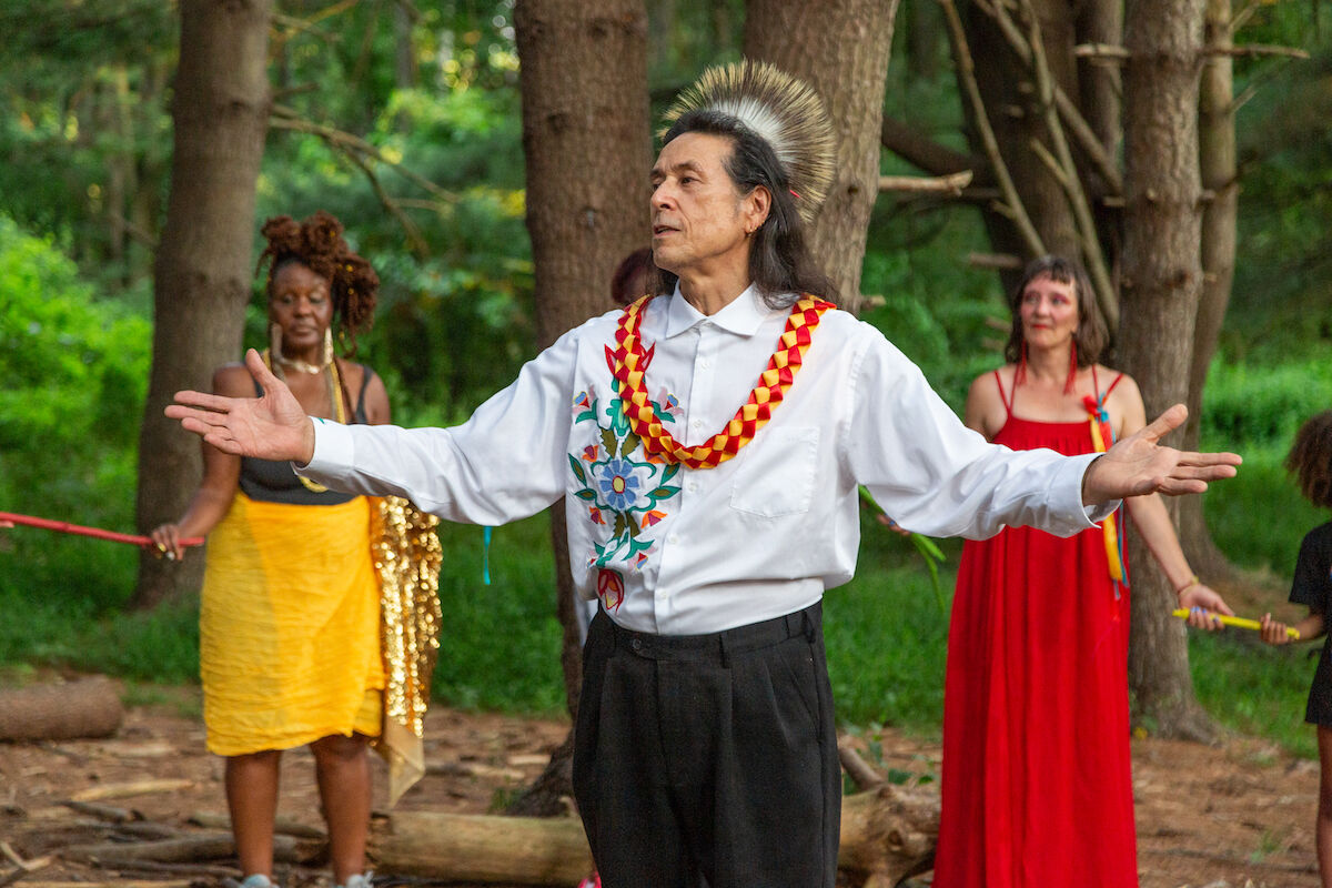 Three figures in the frame, one man in the center flagged by two women behind him. The man's arms are outstretched.