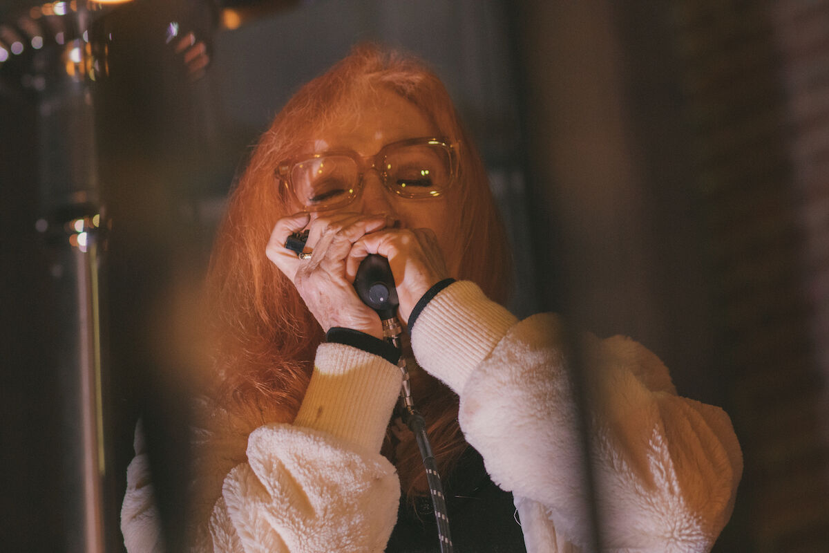 A performer with red hair and eyeglasses plays her harmonica. She's wearing a warm white, fuzzy jacket. A heating lamp keeps her warm in the background.