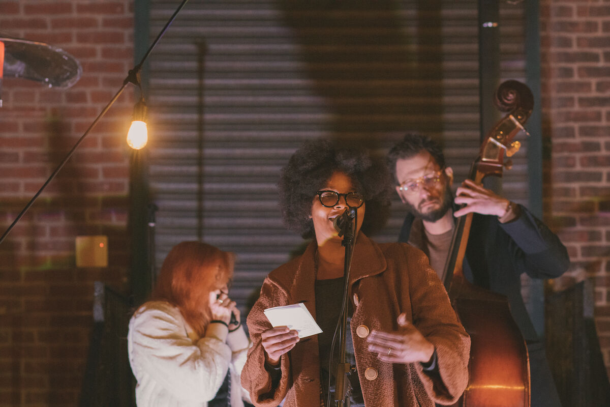 In the foreground, Yolanda Wisher stands in front of a microphone reading notes of a piece of paper in her right hand. Behind her, two performers, one man holding an upright bass and a woman with red hair and a white jacket plays the harmonica.