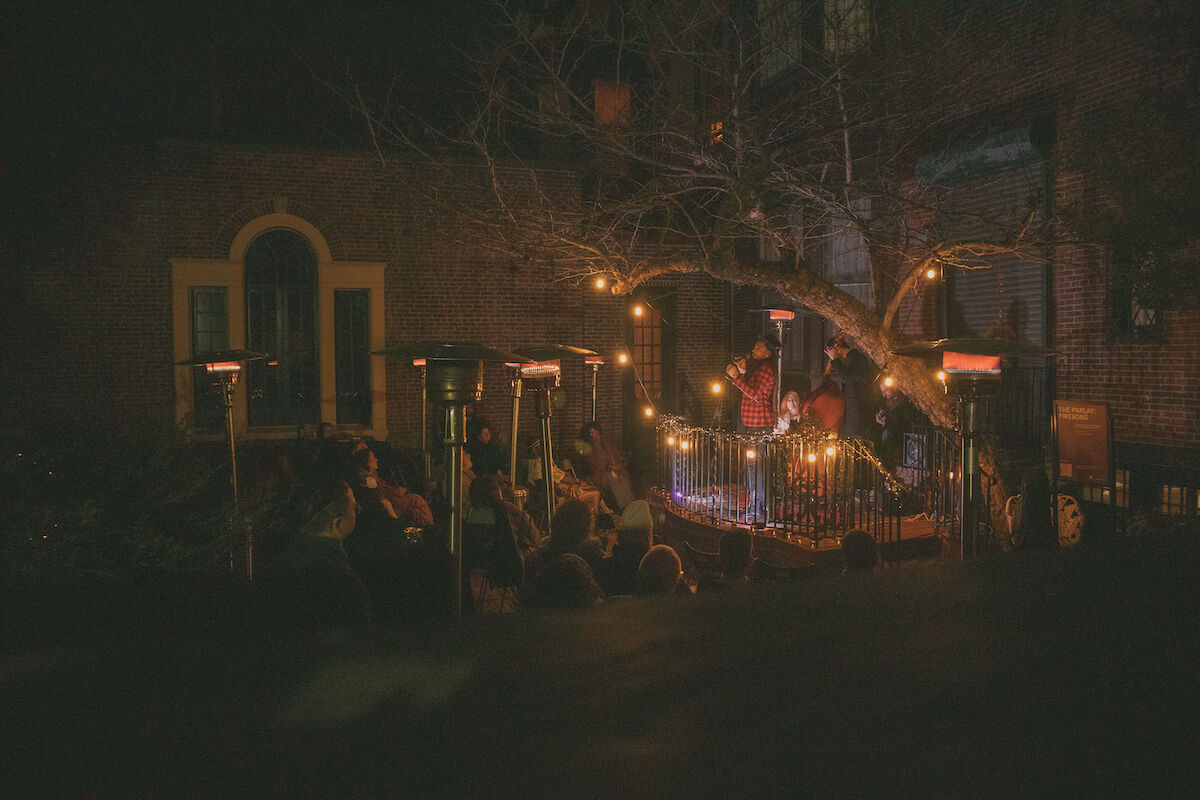 A warm scene after sunset in the backyard of the Rosenbach museum. A large tree with bare branches overhangs on the stage. To the right the performers stand on an elevated stoop, on the left the crowd is warmed by heat lamps.