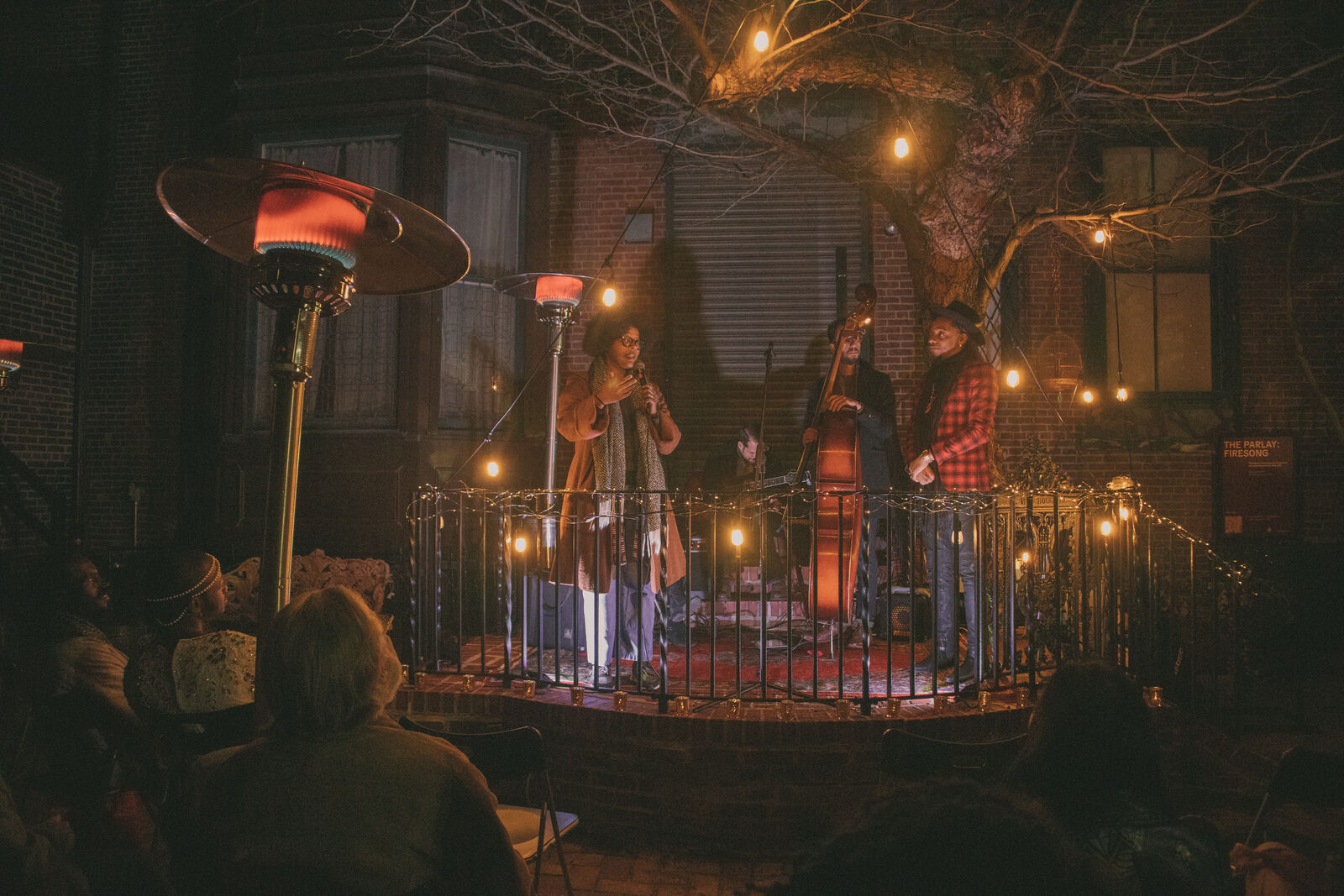 A group of three performers stand on an elevated platform, surrounded by warm fairy lights. In the foreground a crowd watches on. One performer is holding an upright bass, while flanked by two others holding microphones.