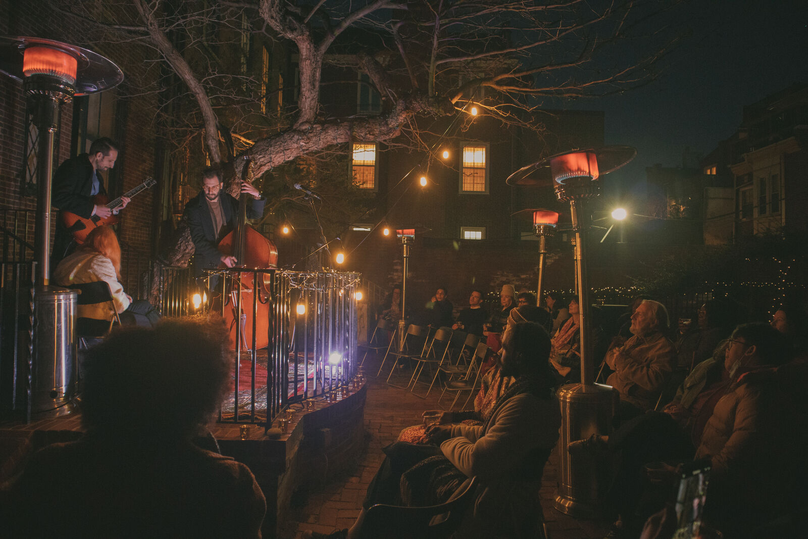 Set at night, on the left musicians perform on top of an elevated stoop surrounded by heat lamps. On the right the crowd looks on, bathed in a warm orange light.