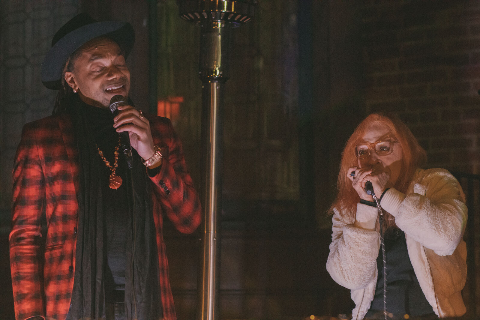 Two performers at night. On the left a male singer holds a microphone; he is clad in a plaid red and black blazer and wearing a fedora. On the right, a female musician plays the harmonic. She has eyeglasses, red hair and wears a white fuzzy coat.