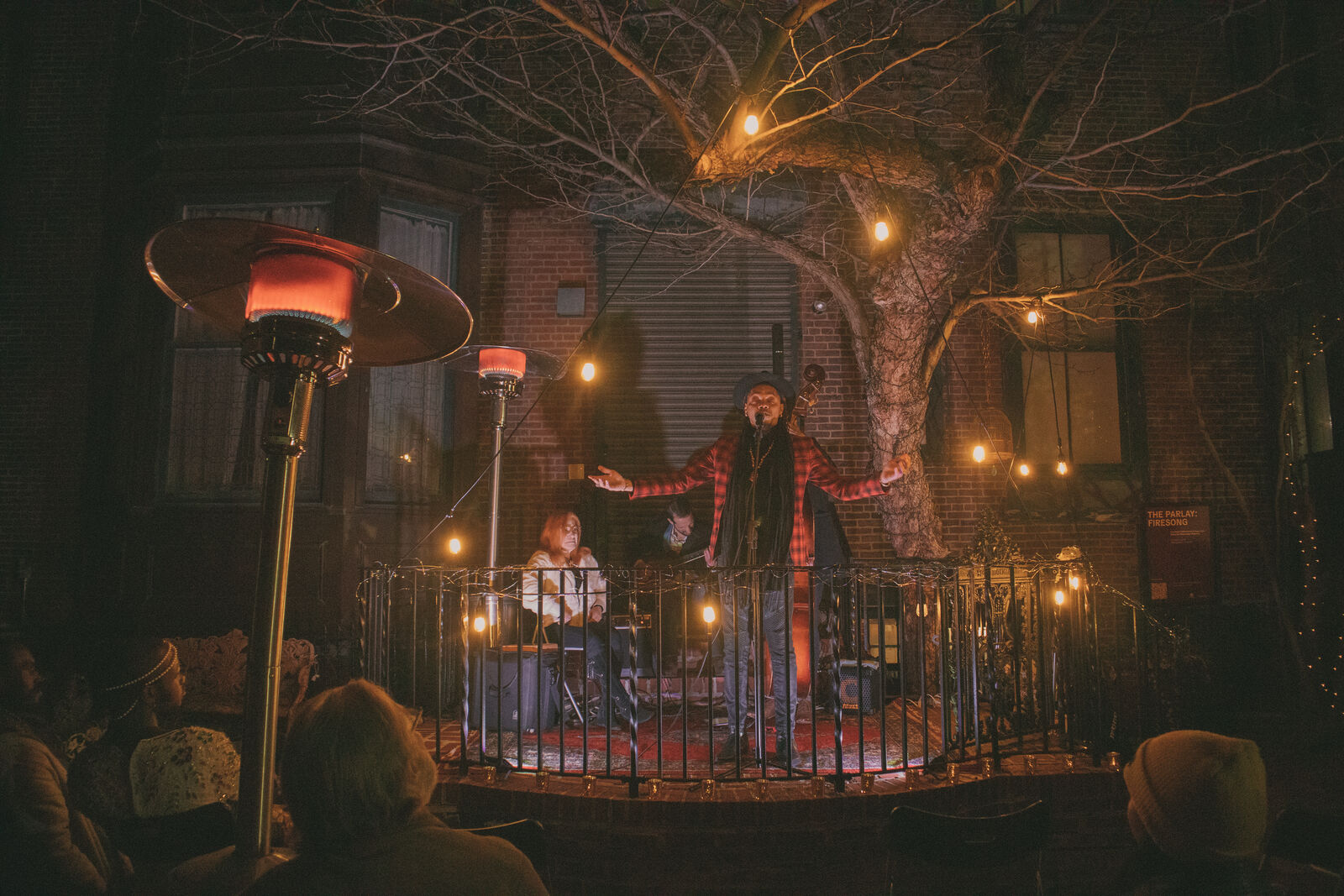 At the center, a male performer stands with arms outstretched, he wears a red and black blazer and a black fedora. Warm fairy lights illuminate the scene and hang from a large bare tree in the background. Heat lamps surround the crowd, keeping them warm in the foreground.