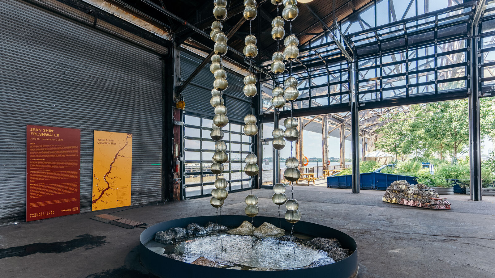 In an open air pier, bulbous glass vessels are suspended in the air from top to bottom. There are four rows of glass vessels, a few contain live mussels that are filtering water in real time. Below the suspended vessels is a shallow fountain basin filled with water trickling down from above, which has been filtered by the mussels. In the basin are pearl blankets in various mounds, the pearls range from snowy white pearlescent to the darker outer shell color of mussels. The pearl blankets were hand sewn and shimmer with each drop of water that comes into contact with them.