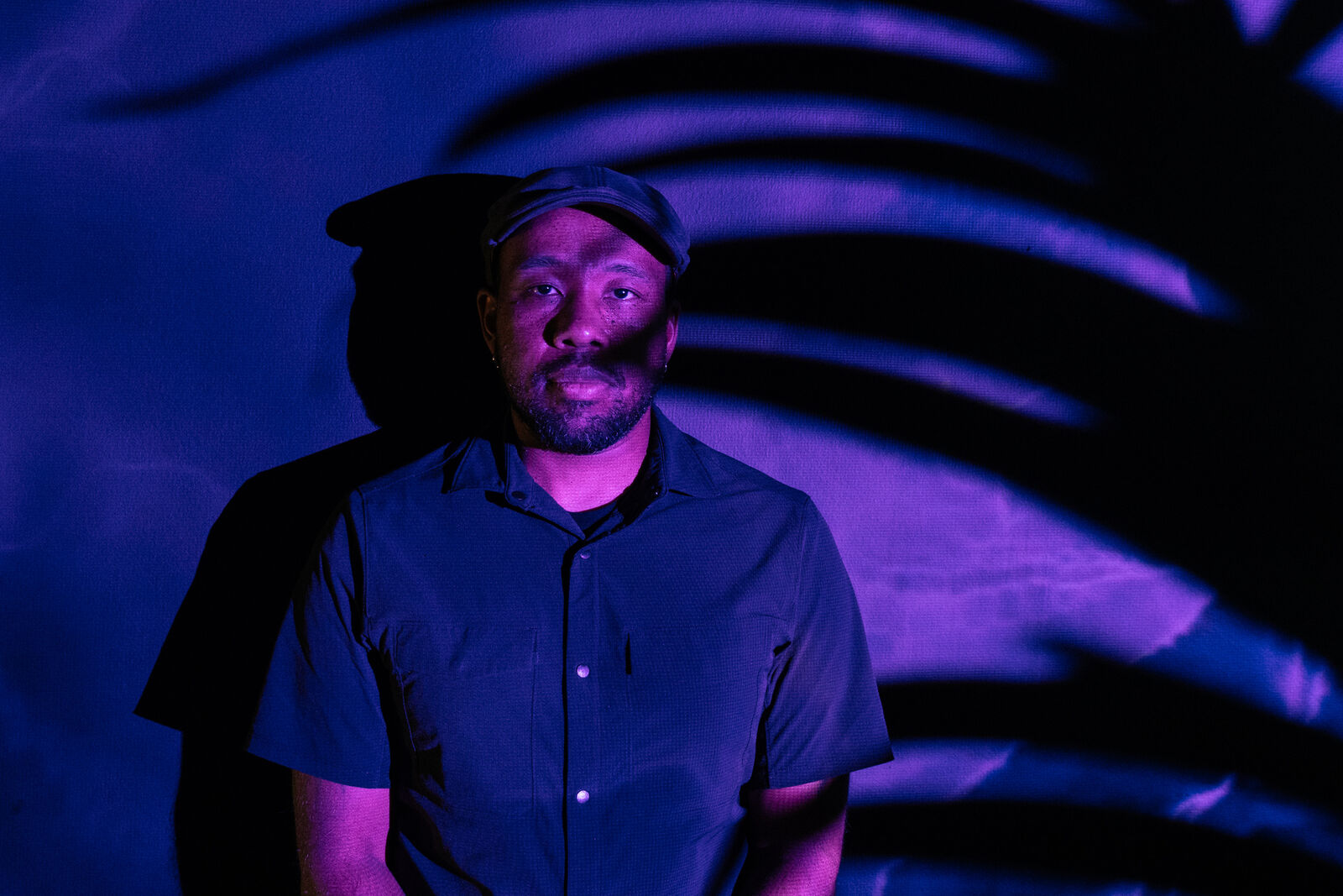 Artist Gralin Hughes, Jr. set against a wall lit by blue and purple lights. A dark shadow from my palm plant covers the right hand side of the image. Hughes wears a cap and short sleeved shirt.