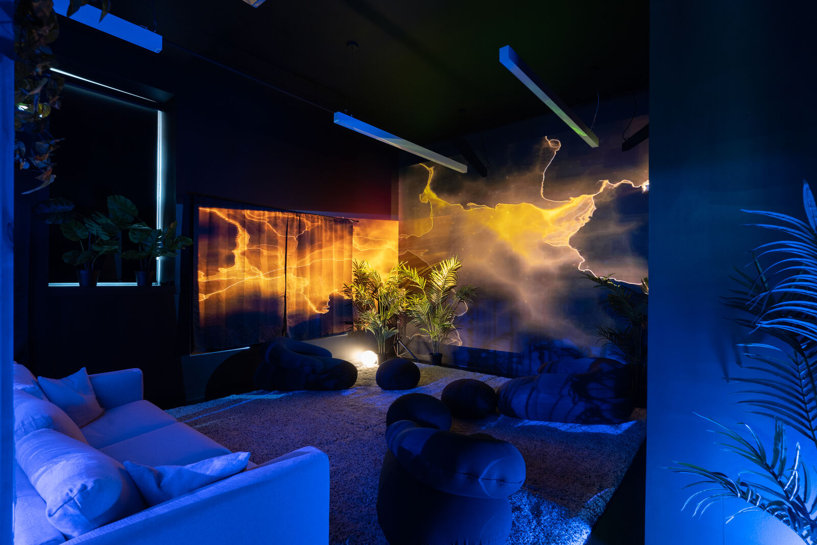 Interior of Supine Horizons exhibition space. Light and visuals are projects onto the walls on the right-hand side, colors are orange and blue. A large white couch is on the left-hand side, smaller dark round cushions are placed in the middle of the room. The leaves of a palm plant peek out on the right corner of the frame.