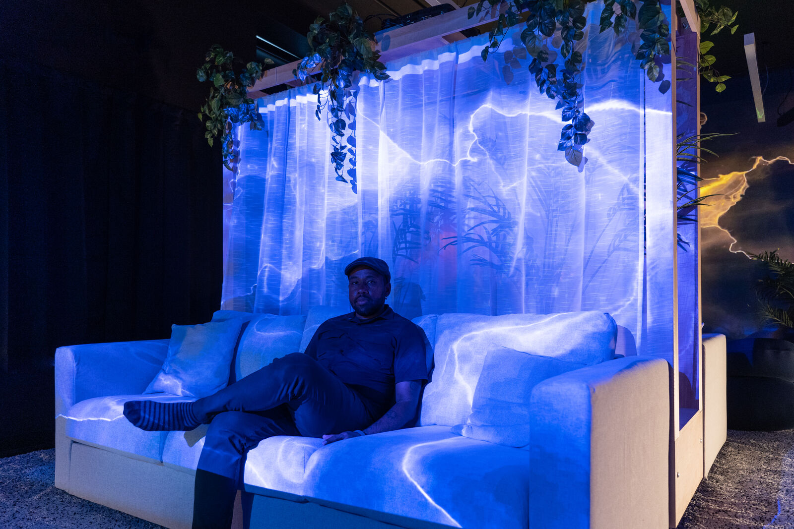 Artist Gralin Hughes, Jr. sits on a white couch, blue and white light surrounds him. A gauzy backdrop is set behind him.
